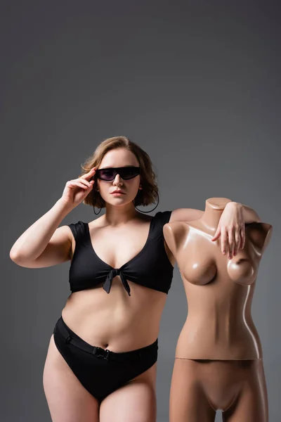 Overweight young woman in swimsuit adjusting sunglasses while posing with plastic mannequin isolated on grey — Stock Photo