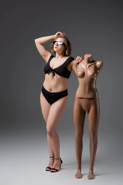 Pleine longueur de jeune femme en surpoids dans des lunettes de soleil posant avec mannequin en plastique sur gris — Photo de stock