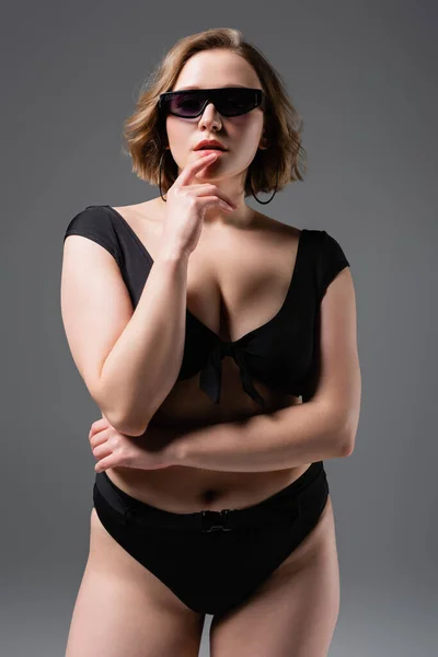 Übergewichtige junge Frau in Sonnenbrille und schwarzem Badeanzug posiert isoliert auf grau — Stockfoto
