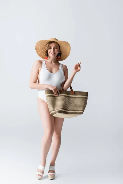 Longitud completa de mujer con sobrepeso y sonrisa en sombrero de paja y traje de baño de pie con bolsa en blanco - foto de stock