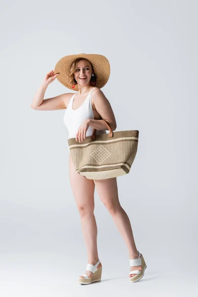 Piena lunghezza di donna sovrappeso e gioiosa in cappello di paglia e costume da bagno in piedi con borsa su bianco — Foto stock