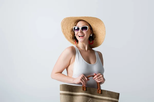 Избыточный вес и веселая женщина в соломенной шляпе, солнцезащитных очках и купальнике, стоящих с мешком изолированы на белом — стоковое фото