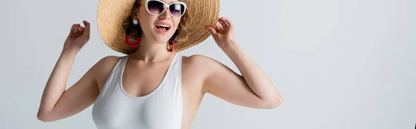 Mujer con sobrepeso y alegre en gafas de sol y traje de baño ajustando sombrero de paja aislado en blanco, bandera - foto de stock