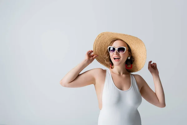 Donna sovrappeso e gioioso in occhiali da sole e costume da bagno regolazione cappello di paglia isolato su bianco — Foto stock