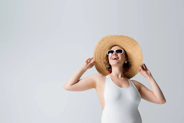 Mujer con sobrepeso y feliz en gafas de sol y traje de baño ajustando sombrero de paja aislado en blanco - foto de stock