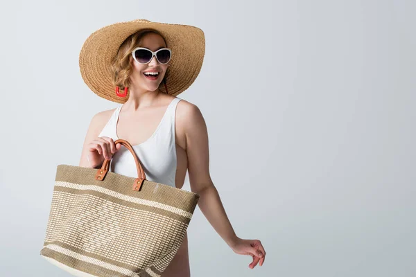 Избыточный вес и веселая женщина в соломенной шляпе, солнцезащитных очках и купальнике держащий сумку изолированы на белом — стоковое фото