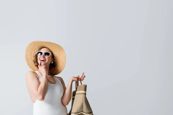 Overweight and cheerful woman in straw hat, sunglasses and swimsuit holding bag isolated on white — Stock Photo