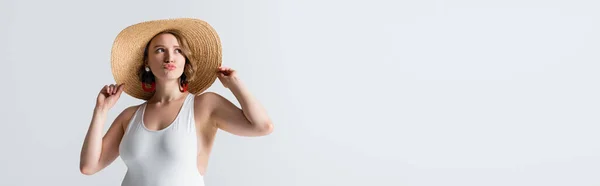 Mulher jovem com sobrepeso em brincos e maiô ajustando chapéu de palha e lábios bebendo isolado em branco, banner — Fotografia de Stock