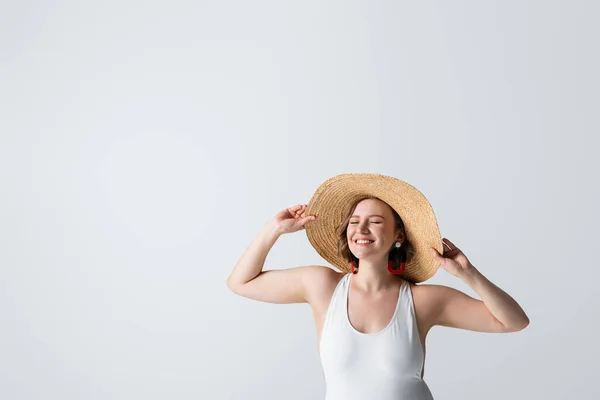 Sobrepeso y mujer complacida en pendientes y traje de baño ajustando sombrero de paja aislado en blanco - foto de stock