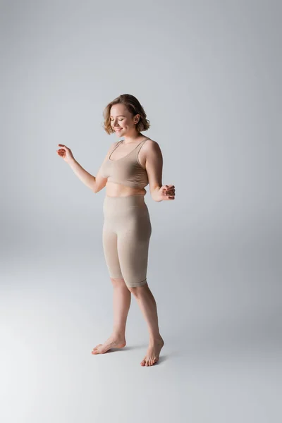 Повна довжина зайвої ваги і задоволена жінка в нижній білизні позує на сірому — стокове фото