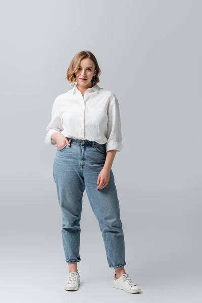 Piena lunghezza di sovrappeso giovane donna in jeans in posa con mano in tasca su grigio — Foto stock