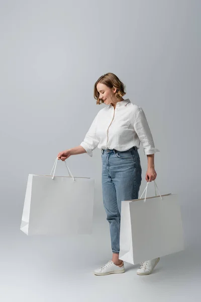 Счастливая толстая женщина в джинсах и белой рубашке, держащая сумки для покупок на серой — стоковое фото