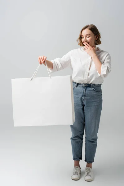 Full length of excited overweight woman in jeans and white shirt holding shopping bag on grey — Stock Photo