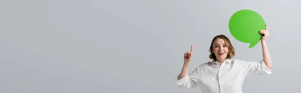 Happy overweight woman in white shirt holding green speech bubble and pointing with finger isolated on grey, banner — Stock Photo