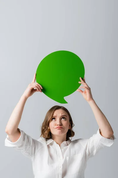 Displeased overweight woman in white shirt holding green speech bubble above head isolated on grey — Stock Photo