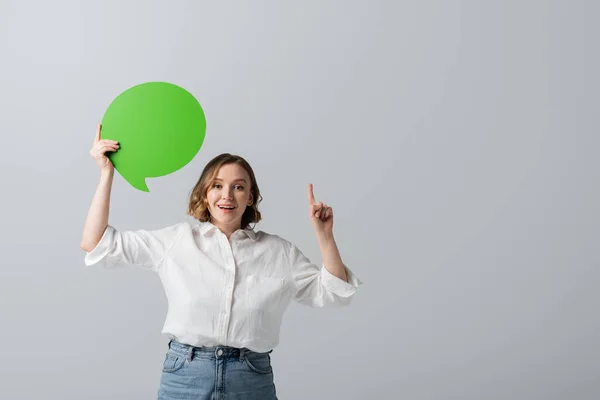 Glücklich übergewichtige Frau im weißen Hemd mit grüner Sprechblase und Zeigefinger isoliert auf grau — Stockfoto