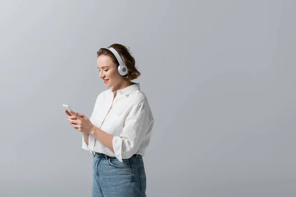 Sonriente mujer de tamaño grande en auriculares inalámbricos escuchando música y sosteniendo el teléfono inteligente aislado en gris - foto de stock