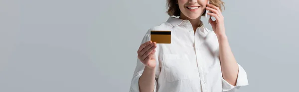 Partial view of plus size woman holding credit card and talking on smartphone isolated on grey, banner — Stock Photo
