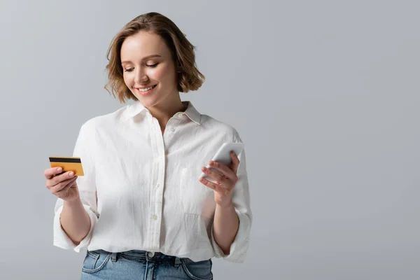 Alegre más tamaño mujer celebración de tarjeta de crédito y teléfono inteligente aislado en gris - foto de stock