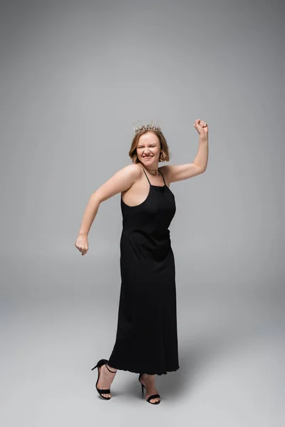 Full length of happy plus size woman in black slip dress and crown on grey — Stock Photo