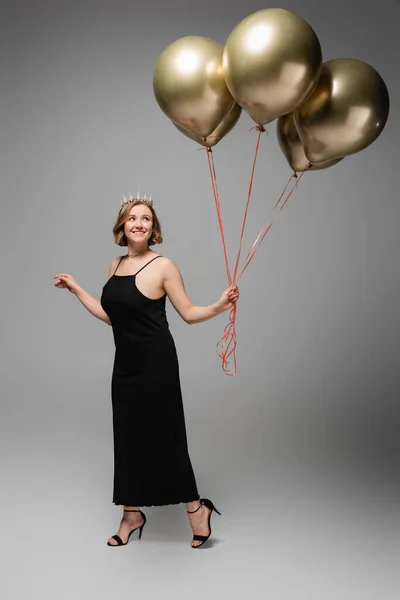 Longitud completa de la mujer feliz más tamaño en vestido de deslizamiento negro y corona con globos de oro en gris - foto de stock