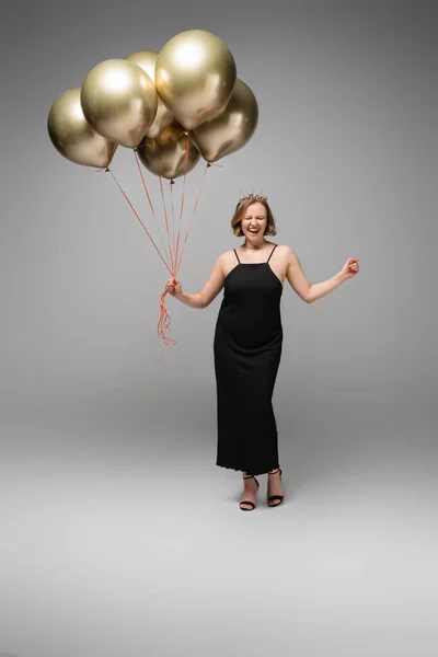 Volle Länge der aufgeregten Plus-Size-Frau in schwarzem Slip-Kleid und Krone mit goldenen Luftballons auf grau — Stockfoto