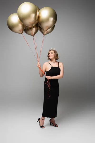 Longueur totale de souriant plus taille femme en robe noire glissante et couronne tenant des ballons dorés sur gris — Photo de stock