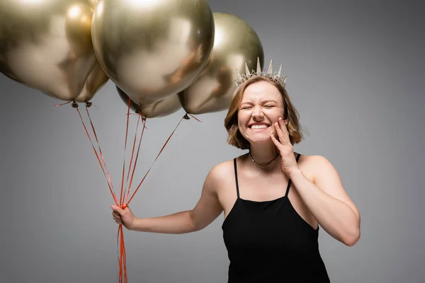 Счастливая plus size женщина в черном платье и короне с золотыми воздушными шариками на сером — стоковое фото