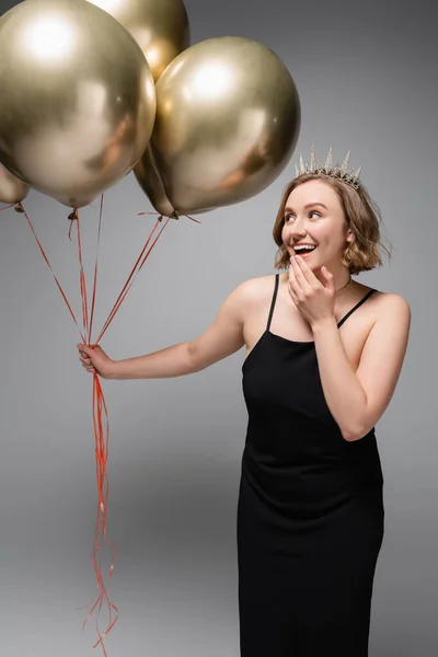 Aufgeregte Plus-Size-Frau in schwarzem Slip-Kleid und Krone mit goldenen Luftballons auf grau — Stockfoto