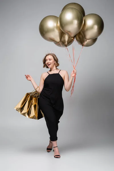 Full length of happy plus size woman in slip dress and crown holding golden balloons and shopping bags on grey — Stock Photo