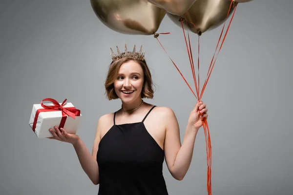 Happy plus size woman in slip dress and crown holding golden balloons and present isolated on grey — Stock Photo