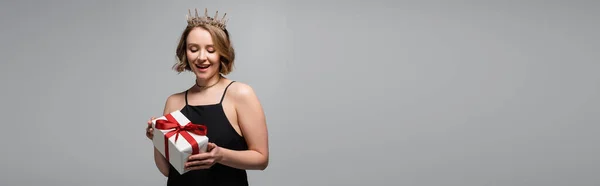 Glückliche Plus-Size-Frau in Slip-Kleid und Krone mit Geschenk isoliert auf grau, Banner — Stockfoto