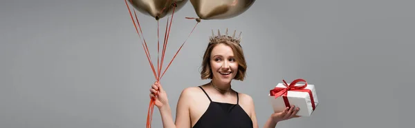 Mujer feliz más tamaño en vestido de deslizamiento y corona con globos de oro y presente aislado en gris, bandera - foto de stock