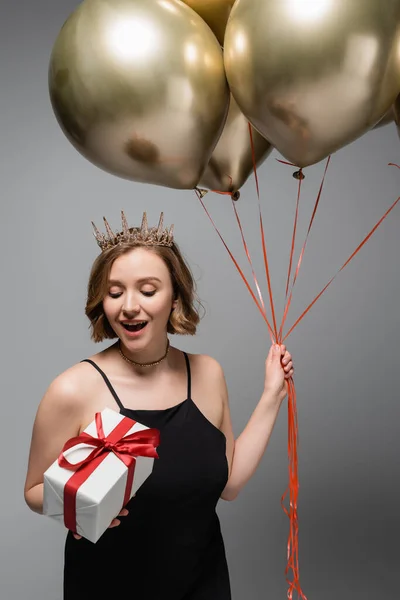 Staunte Plus-Size-Frau in Slip-Kleid und Krone mit goldenen Luftballons und Geschenk isoliert auf grau — Stockfoto