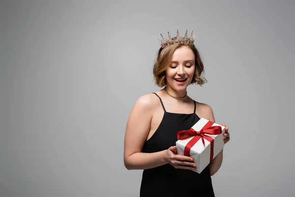 Aufgeregte Plus-Size-Frau in Slip-Kleid und Krone mit Geschenk isoliert auf grau — Stockfoto