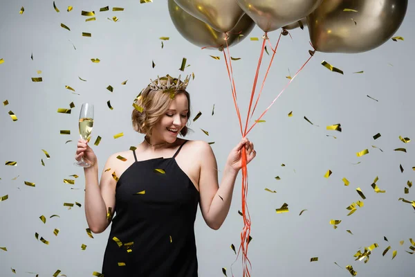 Щаслива жінка плюс розмір в плаття ковзання і корона тримає повітряні кулі і келих шампанського поблизу конфетті на сірому — стокове фото