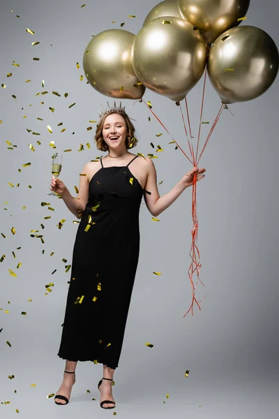 Longitud completa de mujer feliz más tamaño en vestido de deslizamiento y corona celebración globos y copa de champán cerca de confeti en gris - foto de stock