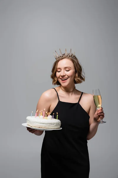 Mujer feliz más tamaño en vestido de deslizamiento y corona celebración pastel de cumpleaños y copa de champán aislado en gris - foto de stock