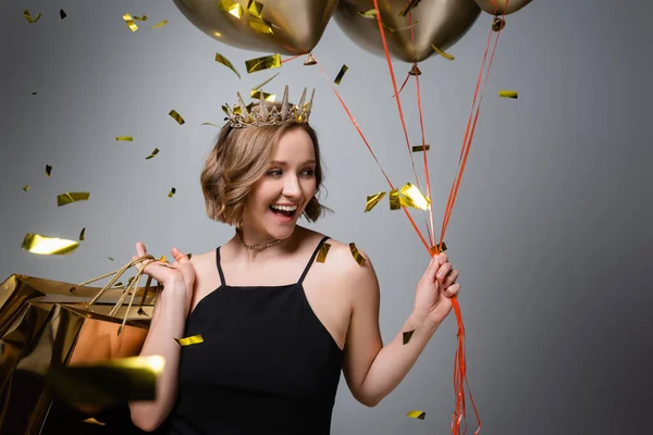 Счастливая plus size женщина в платье и короне, держащая в руках воздушные шарики и сумки для покупок рядом с конфетти на сером — стоковое фото