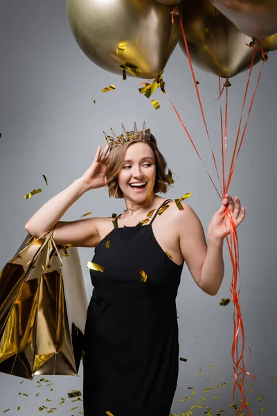 Щаслива жінка плюс розмір в плаття ковзання і корона тримає золоті кулі і сумки біля конфетті на сірому — стокове фото