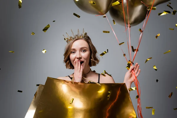 Surprised plus size woman in crown holding balloons and shopping bags near confetti on grey — Stock Photo