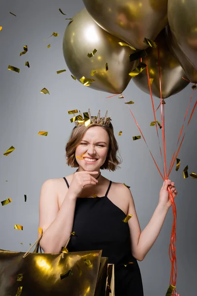 Happy plus size woman in slip dress and crown holding balloons near confetti on grey — Stock Photo