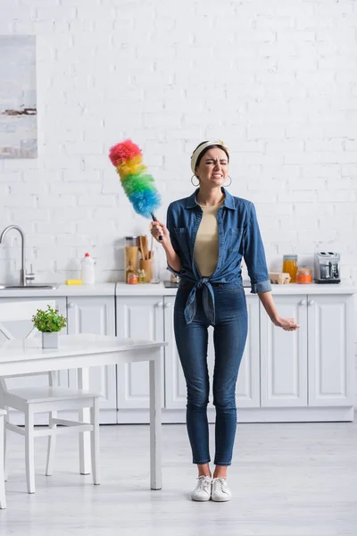 Enojado ama de casa sosteniendo polvo cepillo en cocina - foto de stock
