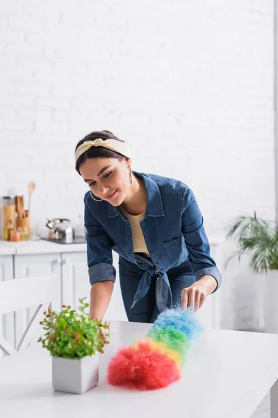 Table de nettoyage femme au foyer avec brosse à poussière près de l'usine floue dans la cuisine — Photo de stock