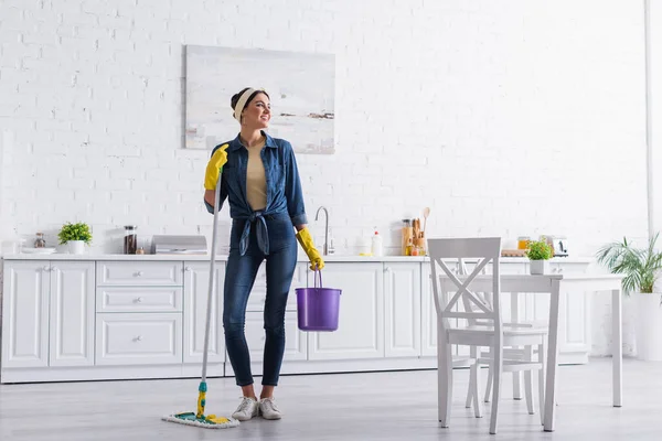 Positivo dona de casa segurando balde e esfregona na cozinha — Fotografia de Stock