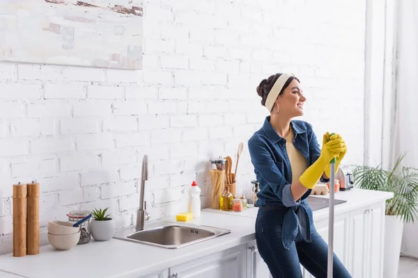 Giovane donna sorridente con scopa in piedi vicino al piano di lavoro della cucina — Foto stock