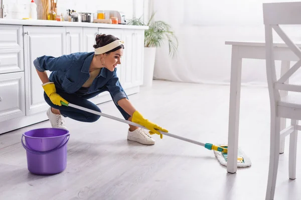 Femme au foyer positive en denim chemise lavage plancher de cuisine — Photo de stock