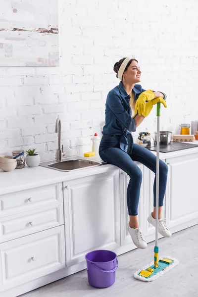 Lächelnde Frau in Gummihandschuhen hält Mopp während sie auf der Arbeitsplatte in der Küche sitzt — Stockfoto