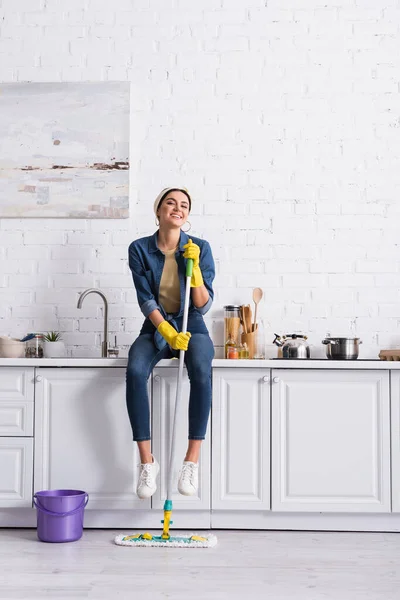 Femme au foyer souriante avec balai assis sur le plan de travail de la cuisine — Photo de stock