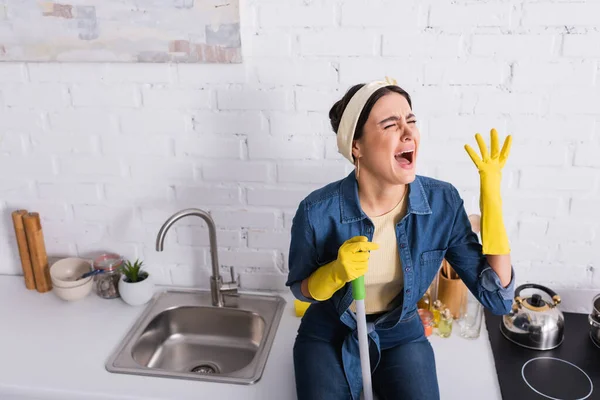 Unzufriedene Hausfrau mit Wischmopp sitzt auf Küchenarbeitsplatte — Stockfoto